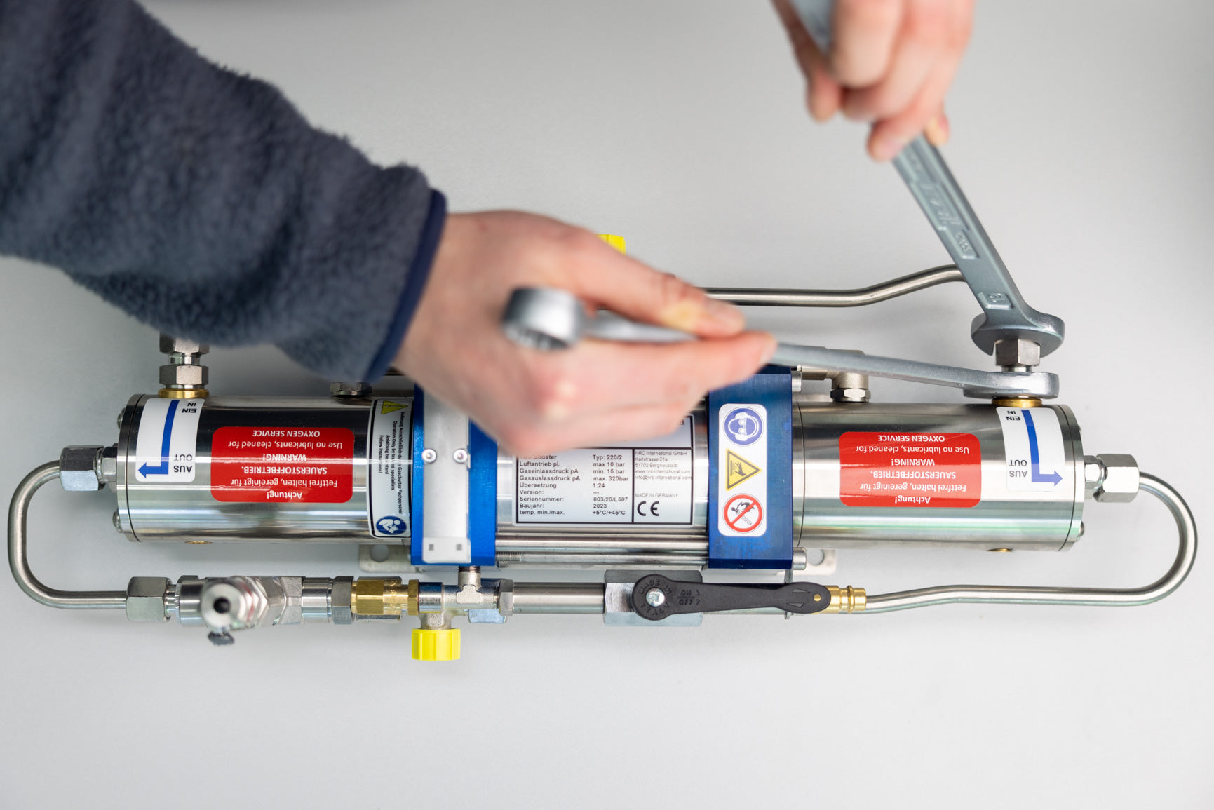 Hands performing maintenance on a high-pressure TEC gas booster, using wrenches to adjust fittings and components on the stainless-steel unit.