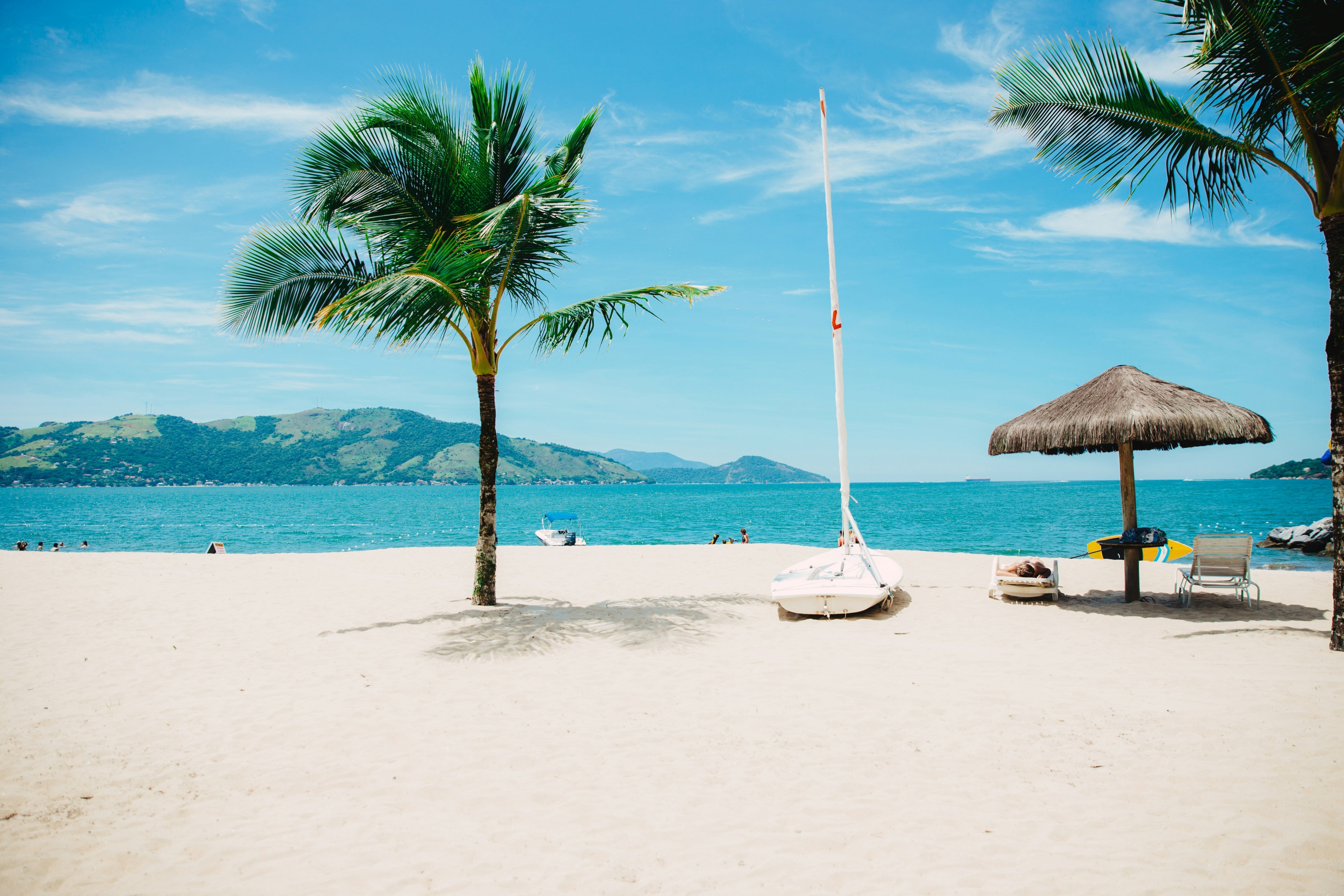 Tropical white sand beach with a palm tree, a small sailboat, and a thatched umbrella against a background of turquoise water and hills.
