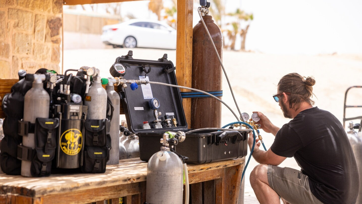 Technician using gas analysis equipment to check mixed-gas cylinders at a dive filling station, with a helium analyzer setup on a workbench.